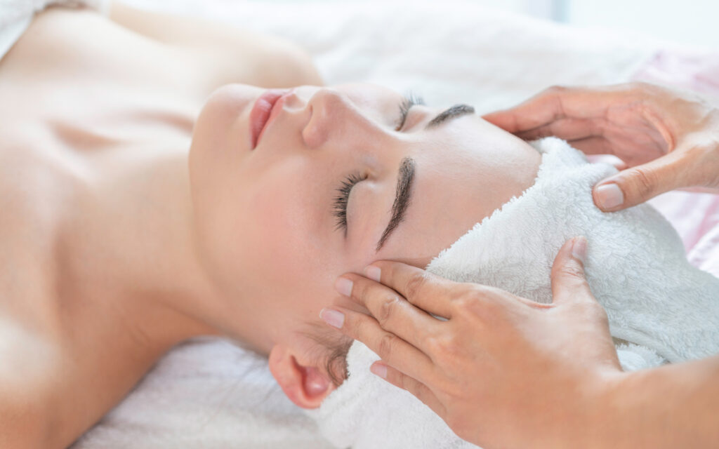 Relaxed woman lying on spa bed for facial and head massage spa treatment by massage therapist in a luxury spa resort. Wellness, stress relief and rejuvenation concept.